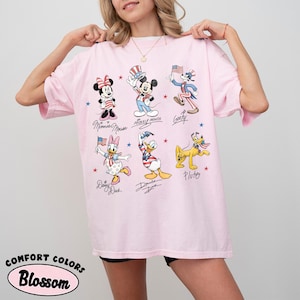May include: A light pink t-shirt featuring a graphic print of Disney characters including Minnie Mouse, Mickey Mouse, Goofy, Daisy Duck, Donald Duck, and Pluto, all dressed in patriotic red, white, and blue outfits with small American flags.