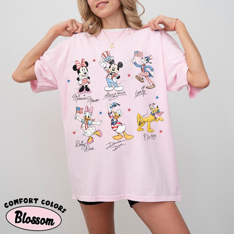 May include: A light pink t-shirt featuring a graphic print of Disney characters including Minnie Mouse, Mickey Mouse, Goofy, Daisy Duck, Donald Duck, and Pluto, all dressed in patriotic red, white, and blue outfits with small American flags.