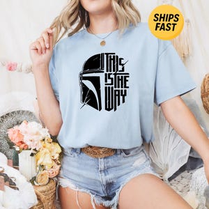 May include: A light blue short-sleeve t-shirt featuring a black graphic print of a helmet silhouette and the text "THIS IS THE WAY" on the front. The shirt has a crew neckline and a relaxed fit, showcasing a bold, stylized design on the chest.