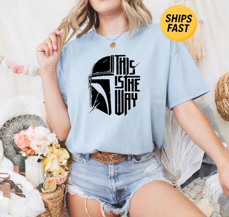 May include: A light blue short-sleeve t-shirt featuring a black graphic print of a helmet silhouette and the text "THIS IS THE WAY" on the front. The shirt has a crew neckline and a relaxed fit, showcasing a bold, stylized design on the chest.
