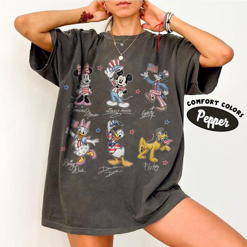 May include: A charcoal gray short-sleeve t-shirt featuring a graphic print of six Disney characters including Minnie Mouse, Mickey Mouse, Goofy, Daisy Duck, Donald Duck, and Pluto, all dressed in patriotic red, white, and blue themed outfits.