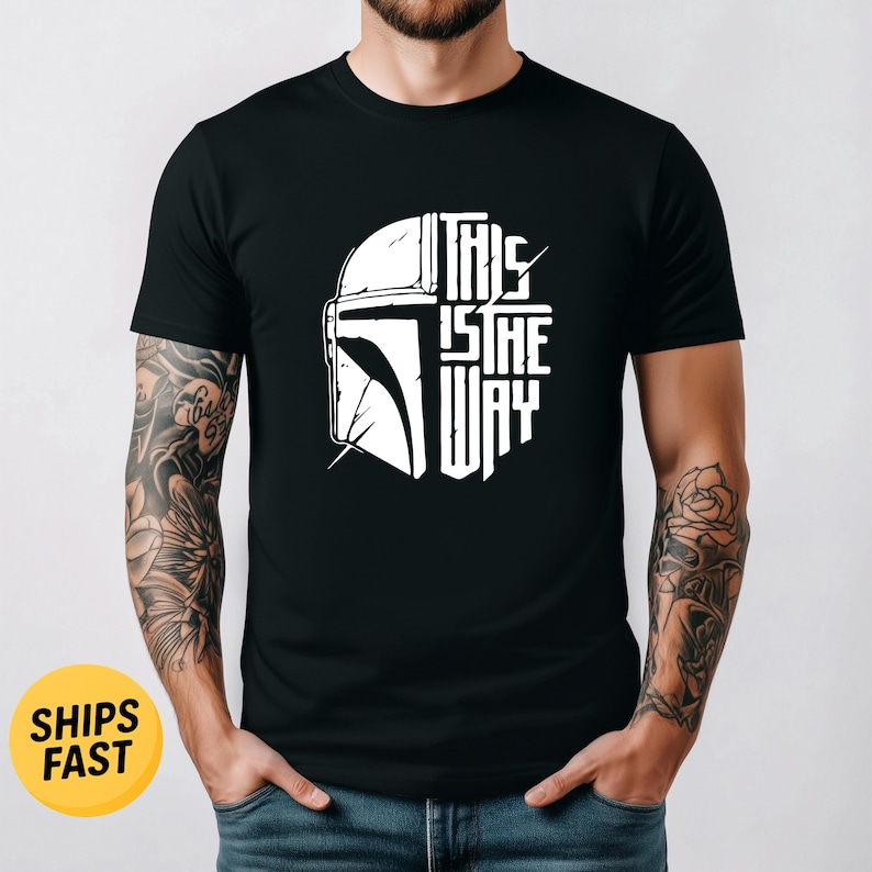 May include: A black short-sleeved t-shirt featuring a white graphic print on the chest. The design displays a stylized helmet silhouette on the left and the text THIS IS THE WAY stacked vertically on the right in a bold, clean font.