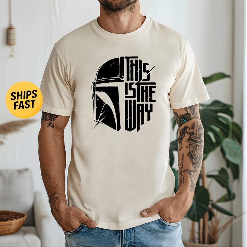 May include: A cream-colored short-sleeve t-shirt featuring a black graphic print on the chest. The design displays a stylized helmet silhouette next to the text "THIS IS THE WAY" arranged in a block formation. The shirt has a crew neckline.