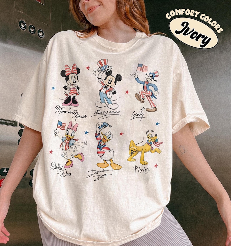 May include: An ivory t-shirt featuring a graphic print of six Disney characters including Minnie Mouse, Mickey Mouse, Goofy, Daisy Duck, Donald Duck, and Pluto, all dressed in patriotic red, white, and blue attire with small stars and text.