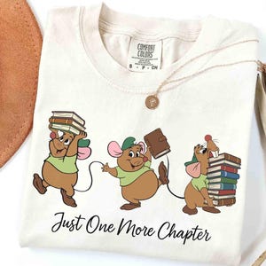 Może przedstawiać: Kremowy t-shirt z grafiką przedstawiającą trzy brązowe myszy w zielonych czapkach i koszulkach, z których każda niesie stosy książek. Poniżej myszy znajduje się czarny napis "Just One More Chapter" wykonany ozdobną czcionką.