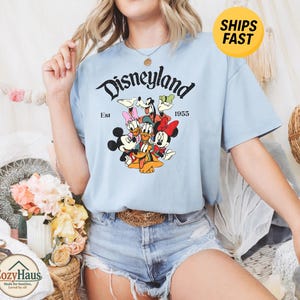 K&ouml;nnte beinhalten: Ein hellblaues Kurzarm-T-Shirt mit einem Grafikdruck von Disney-Figuren, darunter Mickey Maus, Minnie Maus, Donald Duck, Daisy Duck, Goofy und Pluto. Der Text lautet Disneyland Est 1955 in schwarzer Schrift auf dem Shirt.