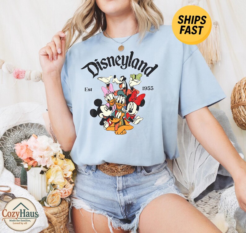 K&ouml;nnte beinhalten: Ein hellblaues Kurzarm-T-Shirt mit einem Grafikdruck von Disney-Figuren, darunter Mickey Maus, Minnie Maus, Donald Duck, Daisy Duck, Goofy und Pluto. Der Text lautet Disneyland Est 1955 in schwarzer Schrift auf dem Shirt.