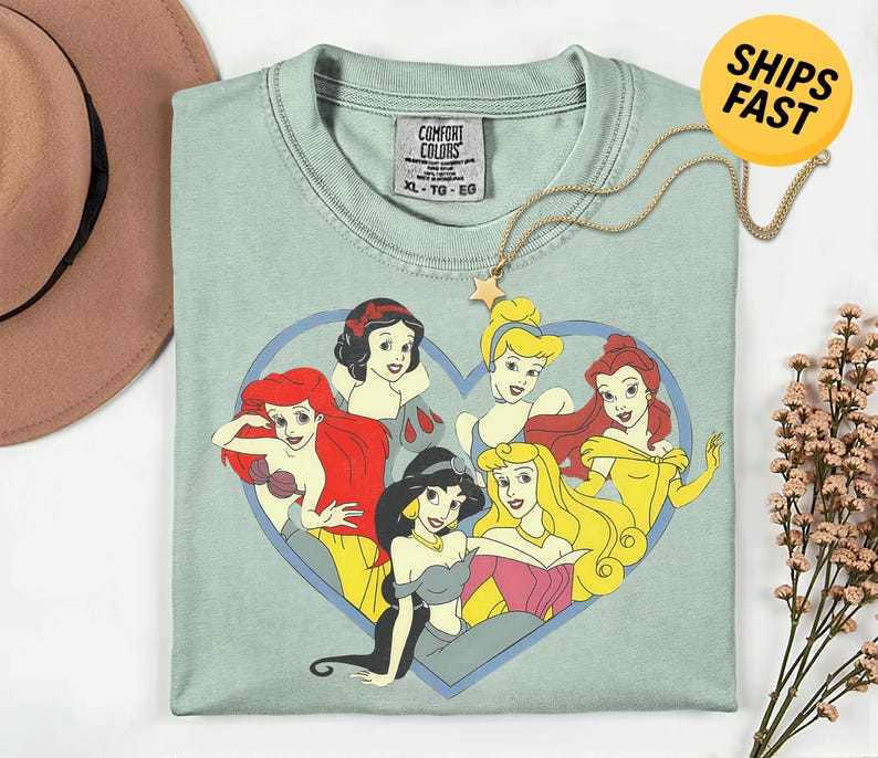 May include: A sage green crew neck t-shirt featuring a graphic print of six animated princesses arranged inside a blue heart outline. The shirt has a Comfort Colors brand label at the inner neckline indicating size XL and is made of 100% cotton.