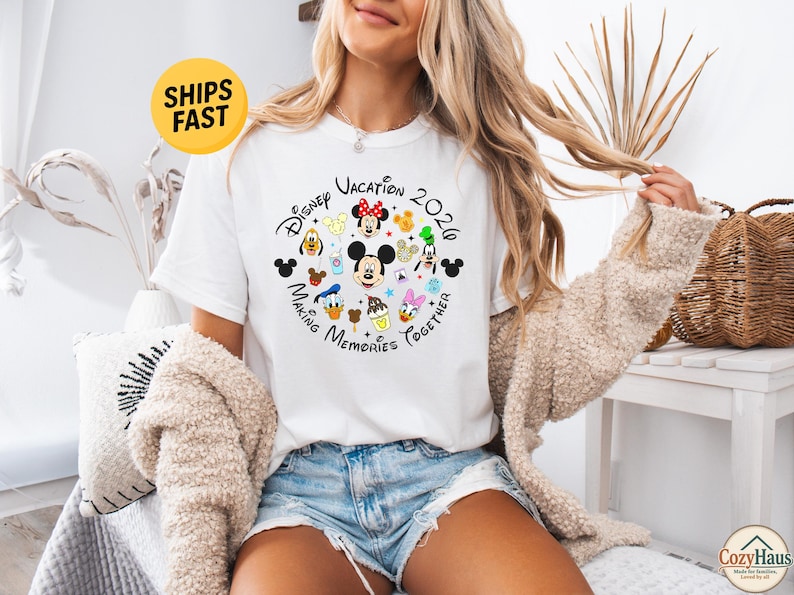 May include: A white short-sleeve t-shirt featuring a circular graphic with the text Disney Vacation 2020 Making Memories Together. The design includes colorful illustrations of Mickey Mouse, Minnie Mouse, Donald Duck, Daisy Duck, Goofy, and Pluto.