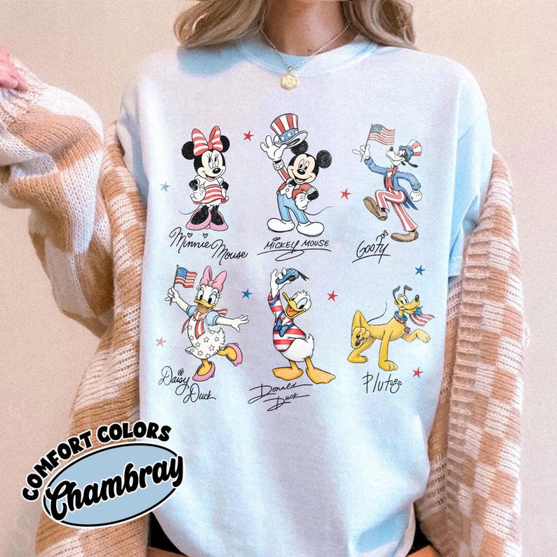May include: A light blue crewneck t-shirt featuring a graphic print of six Disney characters: Minnie Mouse, Mickey Mouse, Goofy, Daisy Duck, Donald Duck, and Pluto. Each character is dressed in patriotic red, white, and blue themed clothing.
