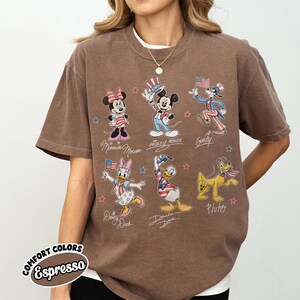 May include: A brown short-sleeve t-shirt featuring a graphic print of six Disney characters including Minnie Mouse, Mickey Mouse, Goofy, Daisy Duck, Donald Duck, and Pluto, all dressed in patriotic red, white, and blue outfits with small stars.