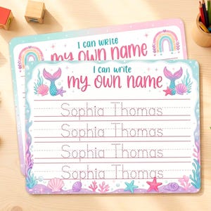 May include: Two rectangular educational writing practice sheets featuring a mermaid theme with pink and blue borders. The text reads I can write my own name, with four lines of dotted text showing the name Sophia Thomas and illustrations of shells.