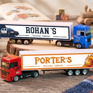 Puede incluir: Dos camiones de juguete con remolque. El camión superior es azul con un remolque blanco etiquetado como "ROHAN'S TRUCKING COMPANY" y un gráfico de coche de policía. El camión inferior es rojo con un remolque blanco etiquetado como "PORTER'S TRUCKING COMPANY" y un gráfico de un balón de baloncesto en llamas.