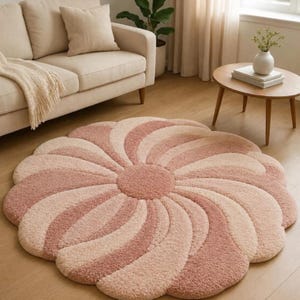 May include: A circular area rug featuring a tufted, high-pile texture with a swirling, petal-like pattern. The design alternates between shades of soft dusty rose and cream, radiating from a solid circular center to create a flower-like shape.