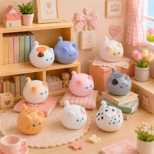 Könnte beinhalten: Eine Sammlung von zehn kleinen, runden, katzenförmigen Figuren in verschiedenen Farben und Mustern, darunter Weiß, Blau, Rosa, Orange und Grau, die auf Holzregalen und Oberflächen neben Miniaturbüchern, einem Haus und Blumendekor stehen.