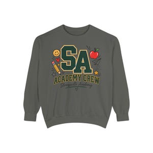 May include: A dark gray crewneck sweatshirt featuring a graphic with the letters SA in large green block font with yellow outlines. Below, the text reads ACADEMY CREW and Strongsville Academy. Icons include a pencil, apple, and smiley face.