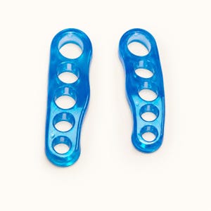 May include: Two blue translucent toe separators made of a smooth, flexible material. Each piece features five circular openings of varying sizes arranged in a vertical row, designed to fit between toes for alignment and spacing purposes.