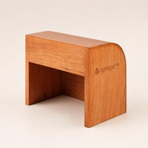 May include: A light brown wooden yoga block with a curved top edge and a flat base. The side of the block features the engraved text iyengar life with a small circular logo. The object has a smooth, natural wood grain finish and a rectangular shape.