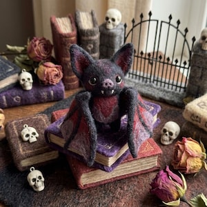 May include: A needle-felted bat figurine with dark gray and deep red fur, large ears, and two small white fangs. The bat is perched on a stack of small, textured books, surrounded by miniature skull charms and dried roses on a dark surface.
