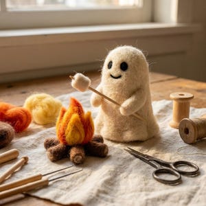 Puede incluir: Una figura de fantasma de lana fieltrada de color crema que sostiene un palo de madera con un pequeño malvavisco blanco, junto a una fogata de lana fieltrada con llamas naranjas y amarillas y troncos marrones, bobinas de madera y tijeras.