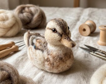 May include: A needle-felted duck figurine crafted from wool, featuring a mottled pattern of cream, tan, and brown fibers. The duck has small, dark, glossy eyes and a textured, rounded body, positioned among various wool roving balls and sewing tools.