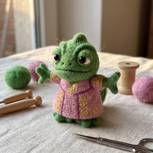 May include: A needle-felted green chameleon figurine wearing a pink jacket with yellow embroidered patterns. The creature has large black eyes and outstretched arms. It is crafted from wool fibers with a textured surface and a soft, rounded appearance.