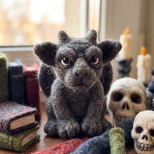 Könnte beinhalten: Eine dunkelgraue, nadelgefilzte Wasserspeier-Figur mit spitzen Ohren, kleinen Hörnern und großen Augen. Die Kreatur sitzt in kauernder Haltung, umgeben von mehreren kleinen gefilzten Büchern, zwei gefilzten Totenköpfen und zwei dünnen Kerzen.