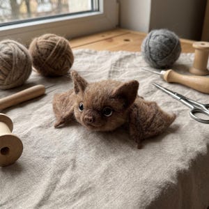 May include: A small, brown, needle-felted bat figurine with textured fur, pointed ears, and dark, glossy eyes. The bat is positioned in the center, surrounded by various crafting supplies including yarn balls, wooden spools, and metal scissors.