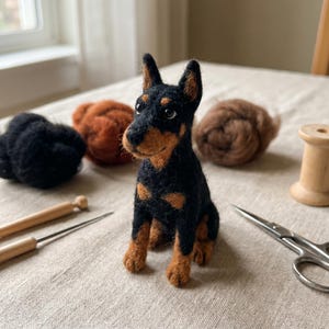 May include: A needle-felted Doberman dog figurine with black and tan fur, featuring pointed ears and dark eyes. The figure is surrounded by loose wool roving in black and brown, a wooden spool, metal scissors, and two felting needles.