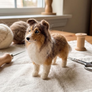 May include: A needle-felted figurine of a Shetland Sheepdog with a sable and white coat. The dog has pointed ears, a black nose, and dark eyes. The textured wool sculpture stands on four legs, capturing the breed's distinct facial markings.