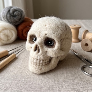 May include: A needle-felted skull sculpture made of off-white wool fiber, featuring dark brown eye sockets with inset black beads, a defined nasal cavity, and a detailed jawline with individual teeth, positioned alongside crafting tools and wool.