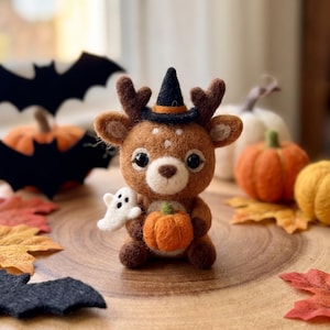 May include: A needle-felted brown deer figurine wearing a black witch hat with an orange band. The deer has large black eyes, white spots, and holds a small orange pumpkin in its paws, with a tiny white ghost figure attached to its side.