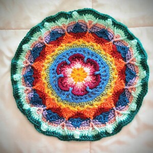 May include: A colourful crocheted mandala with a flower design in the centre. The mandala is made with a variety of colours, including red, orange, yellow, green, blue, and purple.