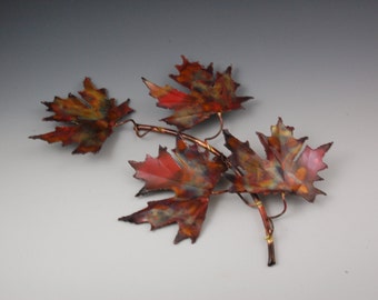 May include: A metal sculpture of maple leaves in shades of red, orange, and green. The leaves are attached to a copper-colored stem. The sculpture is a decorative piece.