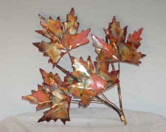 May include: A metal sculpture of maple leaves in shades of red, orange, and gold. The leaves are attached to brown stems, creating a naturalistic, autumnal wall hanging. The sculpture is a decorative piece.