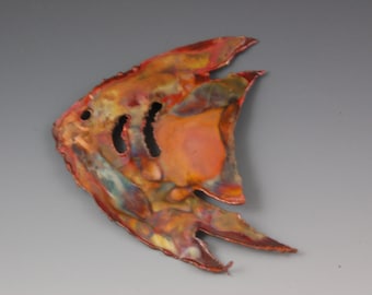 May include: A copper-toned metal fish sculpture with a colorful, textured surface. The fish has two eye holes and two gill slits. The sculpture's colors include shades of red, orange, yellow, and blue.