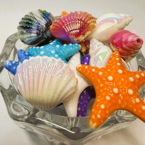 May include: A collection of colorful, glossy, decorative sea-themed beads shaped like seashells and starfish in shades of orange, blue, purple, white, and iridescent pink, contained within a clear, faceted glass bowl with a scalloped edge.