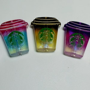 May include: Five small, flat, coffee cup-shaped resin charms in various colors including blue, pink, yellow, purple, and green. Each charm features a glittery finish and a central green circular logo depicting a stylized siren figure.