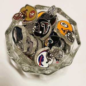 Puede incluir: Un cuenco de cristal transparente lleno de múltiples abalorios de resina con purpurina en forma de cascos de fútbol americano. Cada abalorio muestra los colores y logotipos de varios equipos profesionales, incluyendo diseños en dorado, negro y naranja.