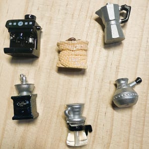 Könnte beinhalten: Eine Sammlung von sechs Miniaturfiguren zum Thema Kaffee, darunter eine schwarze Espressomaschine, ein beigefarbener Jutesack mit Kaffeebohnen, eine silberne Mokkakanne, eine schwarze Kaffeemühle mit der Aufschrift Coffee, ein silberner Cezve und eine Glaskaraffe.