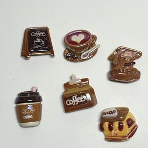 Könnte beinhalten: Ein Set aus sechs kleinen, glänzenden Harz-Anhängern zum Thema Kaffee. Die Teile umfassen ein braunes Aufstellschild mit dem Wort coffee, eine Tasse mit Herz-Latte-Art, eine Kaffeemaschine, einen To-Go-Becher, eine braune Tüte mit der Aufschrift coffee und ein kleines Geschäft.