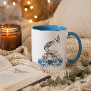May include: A white ceramic mug with a blue interior and handle, featuring a graphic illustration of a tan and white snake coiled among blue flowers, with a small blue butterfly hovering near the snake's head on the front of the mug.