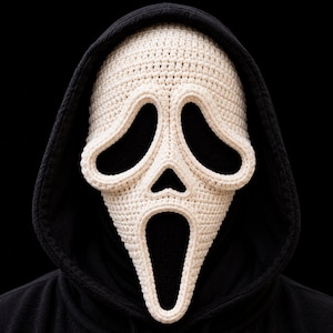 May include: A cream-coloured crocheted mask featuring the iconic elongated ghost face design with large, hollow eye and mouth openings. The mask is worn over a solid black hooded sweatshirt, creating a stark contrast between the light yarn and dark fabric.