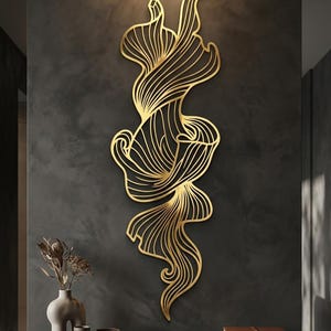 May include: A vertical gold-colored metal wall sculpture featuring an abstract, flowing design with intricate, parallel cut-out lines that create a sense of movement and organic, ribbon-like shapes throughout the entire piece.