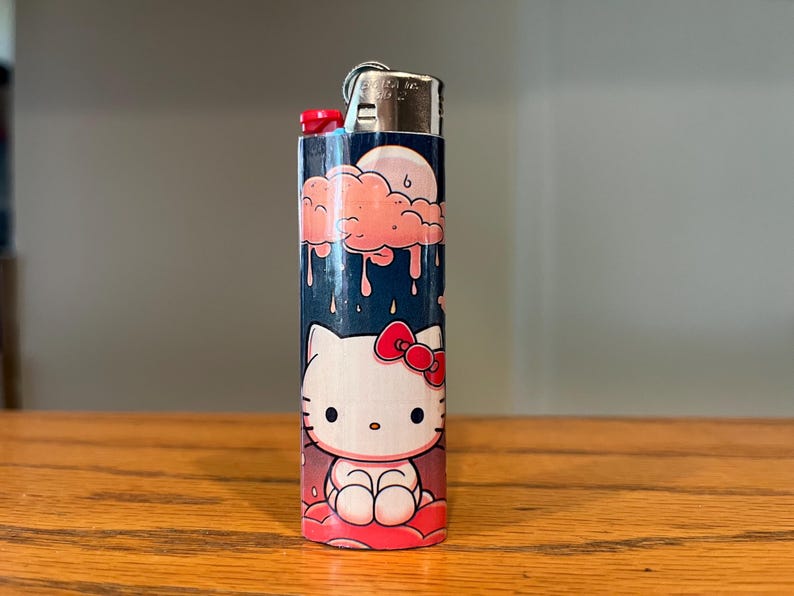May include: A decorative lighter with a Hello Kitty design. The lighter has a blue background with a pink cloud and moon graphic at the top. The bottom of the lighter features a Hello Kitty character with a red bow.