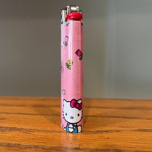 May include: A pink Hello Kitty lighter. The lighter has a red top and is decorated with images of Hello Kitty, flowers, and other small graphics. The lighter is standing upright on a wooden surface.