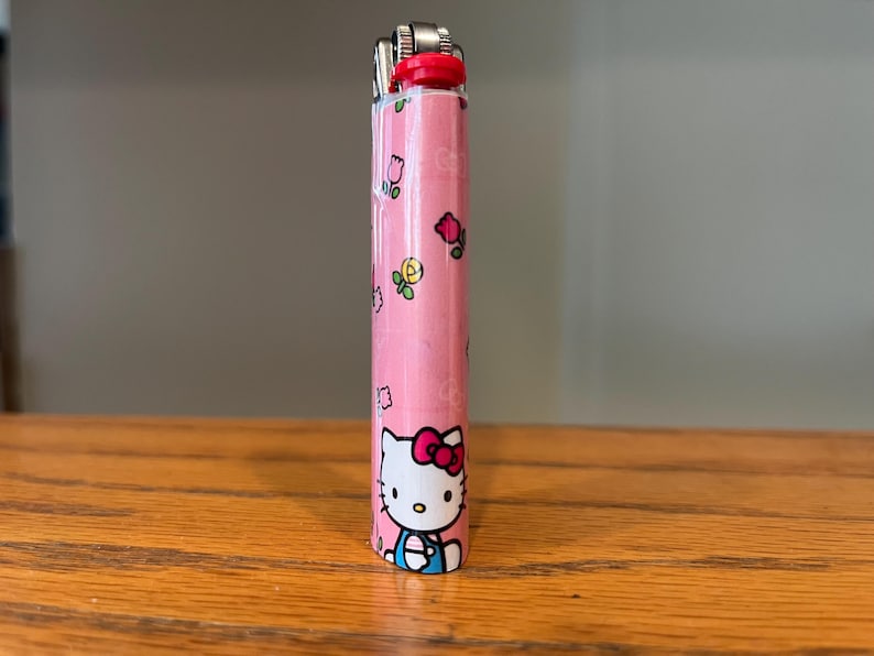 May include: A pink Hello Kitty lighter. The lighter has a red top and is decorated with images of Hello Kitty, flowers, and other small graphics. The lighter is standing upright on a wooden surface.