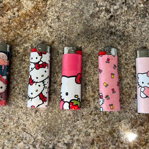 May include: Five Hello Kitty-themed lighters with different designs. One shows a rainy cloud, another has repeating bows, and others feature Hello Kitty with a strawberry, flowers, and a heart. Each lighter has a silver metal top.