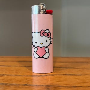 May include: A pink lighter with a Hello Kitty design. The lighter has a silver metal top and a red ignition button. The Hello Kitty graphic shows the character holding a pink heart. The lighter is on a wooden surface.