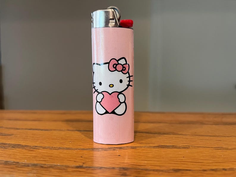May include: A pink lighter with a Hello Kitty design. The lighter has a silver metal top and a red ignition button. The Hello Kitty graphic shows the character holding a pink heart. The lighter is on a wooden surface.